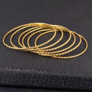 May include: A collection of seven gold-colored bangles. Each bangle has a textured, dotted pattern. The bangles are stacked together, creating a layered effect. The background is a dark, textured surface.