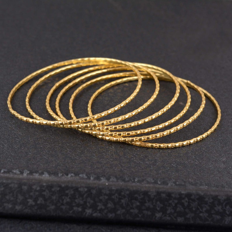 May include: A collection of seven gold-colored bangles. Each bangle has a textured, dotted pattern. The bangles are stacked together, creating a layered effect. The background is a dark, textured surface.