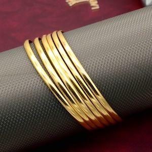 Può includere: Un set di sette braccialetti dorati impilati uno sopra l'altro. I braccialetti sono lisci e lucidi.
