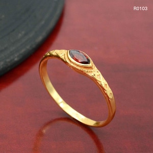 Puede incluir: Un anillo de oro con una piedra preciosa roja oscura con forma de marquesa. La banda y el engaste del anillo presentan intrincados patrones en relieve. El anillo se muestra sobre una superficie roja. El texto "R0103" es visible.