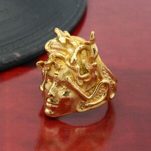 May include: A gold-colored ring with a detailed design of a woman's face with snakes for hair. The ring is made of metal and has a textured surface.