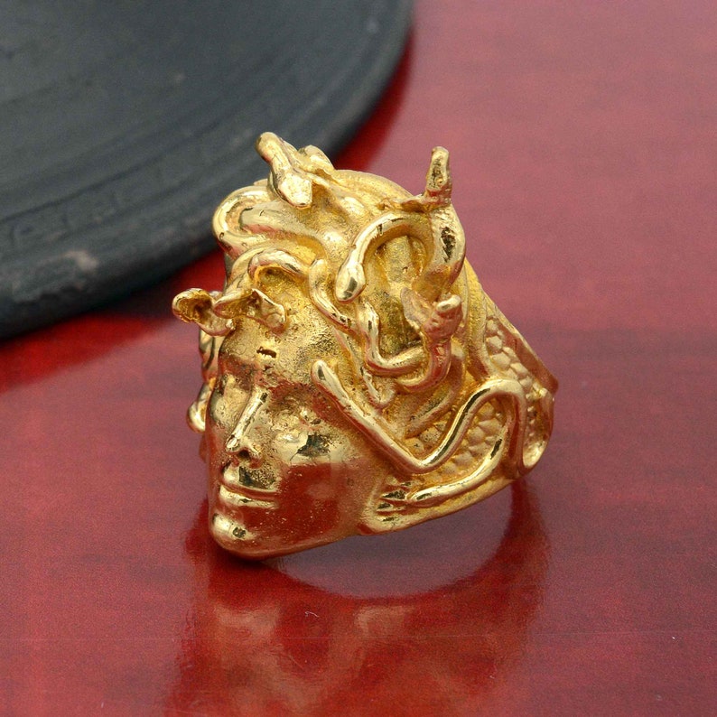May include: A gold-colored ring with a detailed design of a woman's face with snakes for hair. The ring is made of metal and has a textured surface.