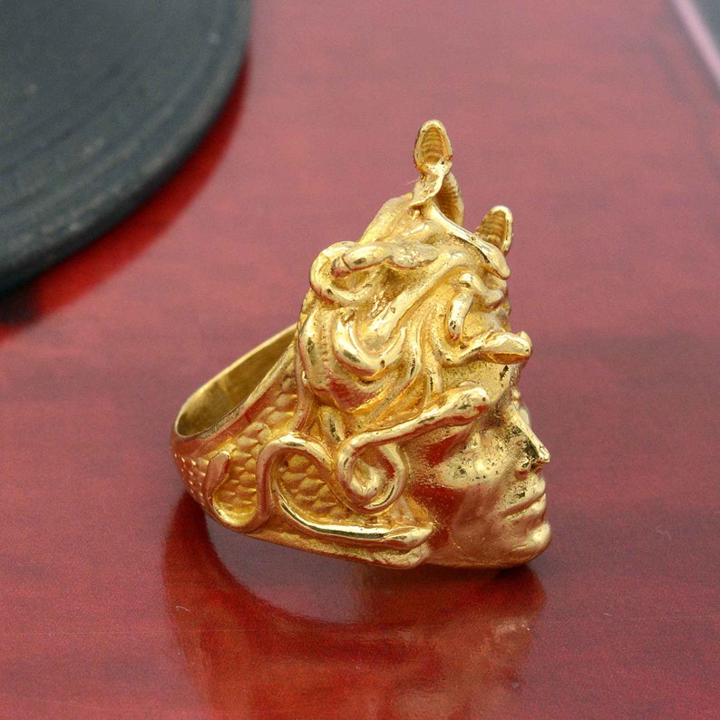 May include: A gold-toned ring featuring a detailed sculpted design of a woman's face with snakes in her hair.