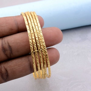 Może przedstawiać: Zestaw sześciu złotych bransoletek z fakturowanym wzorem. Bransoletki są ułożone jedna na drugiej.