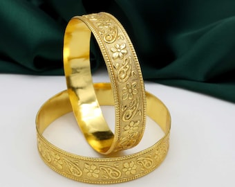 May include: Two gold bangles with intricate floral and paisley designs. The bangles are stacked on top of each other.