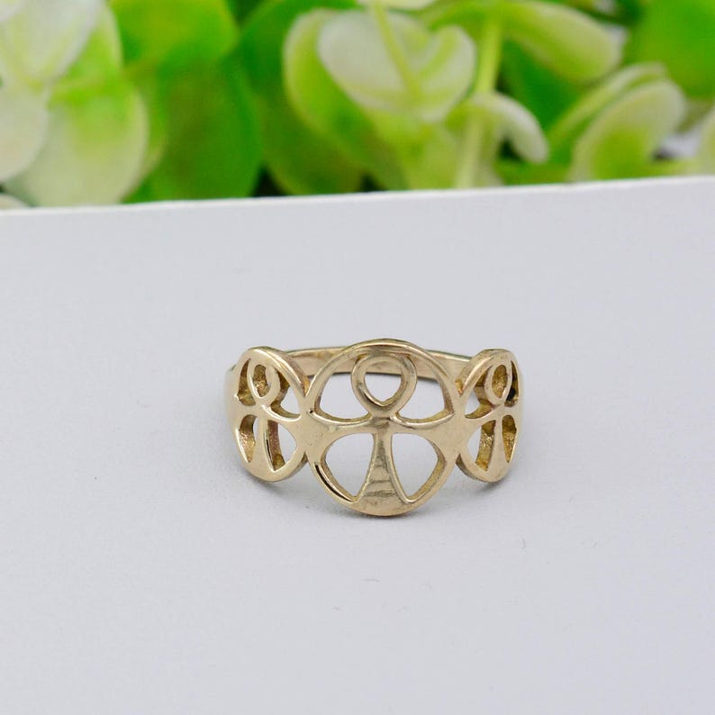 May include: A gold-colored ring with an openwork design featuring an Ankh symbol in the center. The ring has a smooth, polished finish and is set against a white background. The ring's design is symmetrical and detailed.