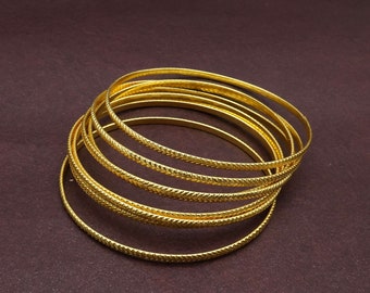 May include: A stack of eight gold-colored bangles with a textured design. The bangles are stacked on top of each other and are arranged in a circular pattern.