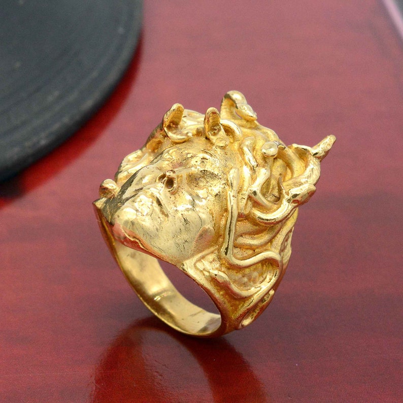 May include: A gold-colored ring with a detailed design of a face with snakes for hair. The ring is on a red surface.