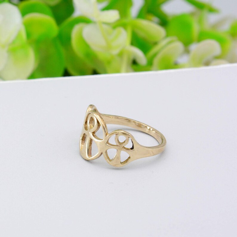 May include: A gold-colored ring with an openwork design. The ring features a stylized floral or geometric pattern. The ring is set against a white background with blurred green foliage in the background.