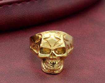 May include: A gold-coloured skull ring with intricate details. The ring is made of metal and features a detailed skull design.