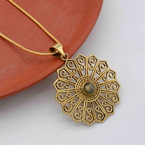 Könnte beinhalten: Eine goldene Halskette mit einem großen, verzierten Anhänger, der ein kreisförmiges Design mit einem zentralen Edelstein aufweist. Der Anhänger hat ein filigranes Muster und ist von einem Ring aus kleineren Kreisen umgeben.