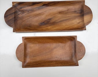 May include: Two rectangular wooden serving trays with rounded handles. The trays are made of a dark brown wood with visible grain patterns. The larger tray is positioned above the smaller one, both resting on a white surface.