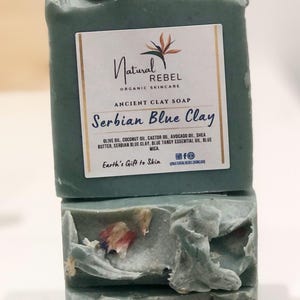 May include: Two bars of blue-green soap with dried flowers embedded in the soap. The soap is labeled "Natural Rebel Organic Skincare Ancient Clay Soap Serbian Blue Clay Olive Oil, Coconut Oil, Castor Oil, Avocado Oil, Shea Butter, Serbian Blue Clay, Blue Tansy Essential Oil, Blue Mica Earth's Gift to Skin"