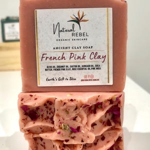 May include: A pink bar of soap with the text "Natural Rebel Organic Skincare Ancient Clay Soap French Pink Clay" and the ingredients listed below. The soap is decorated with pink rose petals.