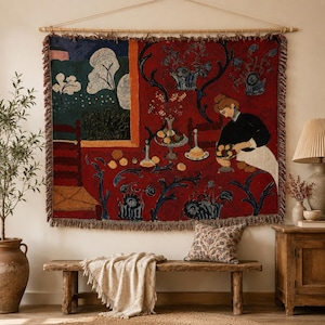 May include: A rectangular woven wall tapestry featuring a vibrant red background with a dark blue floral pattern. The scene depicts a person in a black top and white skirt at a table with fruit, next to a window showing trees and a blue sky.
