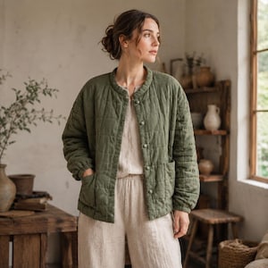 Puede incluir: Una chaqueta acolchada de color verde oliva con cuello redondo, mangas largas con puños elásticos y cierre frontal de botones. La chaqueta incluye dos bolsillos de parche en la parte delantera y un patrón de costuras en forma de cuadrícula.