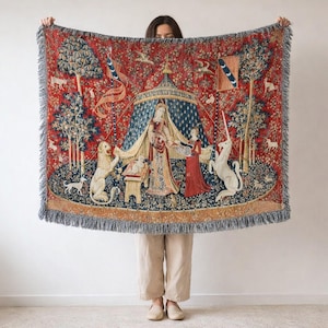 Puede incluir: Una manta tejida con un tapiz que representa una escena medieval con una mujer, un unicornio y un león. La manta tiene un fondo rojo con detalles en azul y beige, y un borde con flecos. El diseño representa una tienda de campaña, árboles y varios animales.