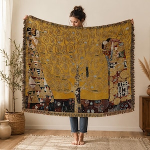 Op de afbeelding: Een geweven plaid met een goudgeel levensboomontwerp met ingewikkelde wervelende takken, geometrische patronen en twee figuren in gewaden. Het rechthoekige textiel heeft een gefranjerde rand en toont een rijk palet van goud, zwart en bruin.