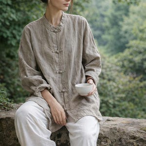 Può includere: Una persona indossa una camicia di lino marrone chiaro con colletto alla coreana e pantaloni bianchi ampi, tenendo in mano una piccola ciotola bianca. La camicia ha chiusure con bottoni. Lo sfondo è verde sfocato, che suggerisce un ambiente esterno.