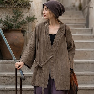 May include: A brown, textured kimono-style jacket with a matching belt, worn over a black top and purple trousers. The jacket has wide sleeves and a relaxed fit. A brown beanie and a patterned bag complete the outfit. The background features stone steps and a large terracotta pot.