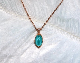 Puede incluir: Un collar de oro rosa con un colgante ovalado de color turquesa. El colgante está enmarcado en un marco dorado decorativo con un pequeño diamante. El collar se muestra sobre un fondo blanco y plumoso.