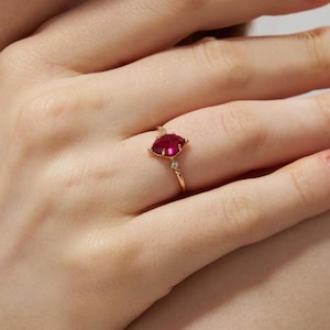 May include: A gold ring with a red gemstone and two smaller white gemstones. The ring is on a person's finger.