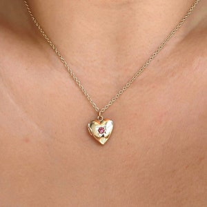 May include: A gold heart-shaped locket necklace with a small red gemstone in the centre.