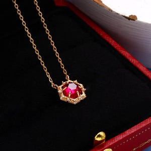 May include: A gold chain necklace with a red gemstone pendant. The pendant is surrounded by smaller clear gemstones and has a detailed design.