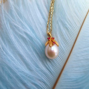 以下が含まれることがあります： 大きな白い真珠のペンダントが付いたゴールドネックレス。ペンダントは小さな赤い宝石と金の葉のアクセントで飾られています。ネックレスは柔らかな青い羽根の背景に飾られています。