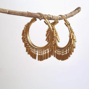 Peut inclure: Une paire de boucles d'oreilles créoles dorées avec une frange de petites barres dorées qui pendent du bas.