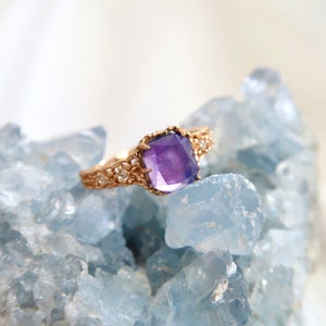 May include: A gold ring with a large, square-shaped amethyst gemstone set in a delicate, ornate design. The ring is resting on a bed of blue crystals.