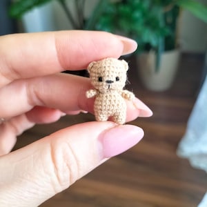 Könnte beinhalten: Ein winziger, handgehäkelter Teddybär in hellbrauner Farbe. Der Bär hat kleine schwarze Augen und eine aufgestickte Nase. Die Arme und Beine sind kurz und stämmig. Der Bär ist etwa 2,5 cm groß, ein Miniatur-Stofftier.
