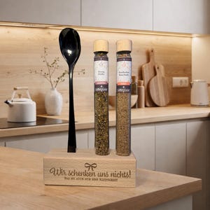 Könnte beinhalten: Ein hölzerner Gewürzständer mit zwei Gewürzgläsern und einem schwarzen Löffel. Die Gläser haben Holzdeckel und Etiketten. Auf dem Ständer steht der Text "Wir schenken uns nichts!" und "Das ist auch nur eine Kleinigkeit".