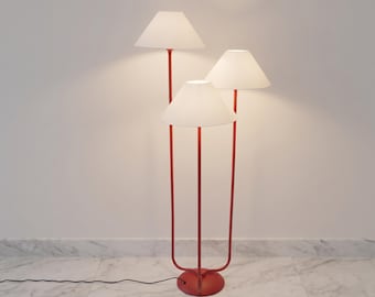 May include: A floor lamp featuring three red metal poles of varying heights, each topped with a white conical lampshade. The poles are connected to a single circular red base, and a black power cord extends from the base along the floor.