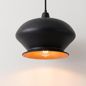 May include: A matte black pendant light fixture with a wide, rounded dome shape. The interior of the shade features a warm, metallic gold finish. A single clear glass light bulb is visible inside, emitting a soft, warm glow from the center.