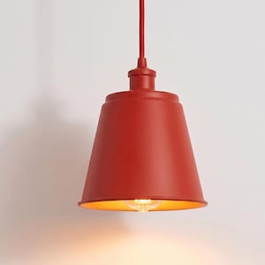 May include: A matte red pendant light fixture with a conical shade and a matching red cord. The interior of the shade is a warm yellow tone, and a single light bulb is visible at the center, emitting a soft glow from the bottom opening.