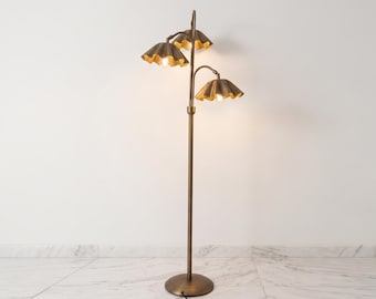 Puede incluir: Una lámpara de pie con un soporte de metal alto y delgado en tono latón y una base circular. La lámpara cuenta con tres brazos ajustables, cada uno terminado en una pantalla festoneada en forma de campana con acabado metálico que emite luz cálida.
