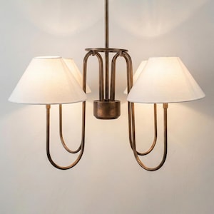 May include: A bronze-colored chandelier with four white lampshades. The light fixtures have a curved, minimalist design. The lampshades are cone-shaped and emit a warm glow. The chandelier is suspended from the ceiling.