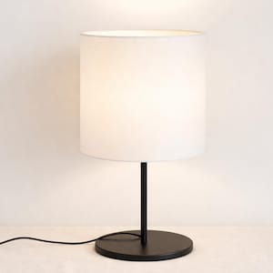 May include: A modern table lamp with a white cylindrical shade and a black base and stem. The lamp is turned on, emitting a soft, warm glow. The lamp's cord is visible, extending to the left. The lamp is a simple, elegant design.
