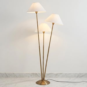 May include: A floor lamp featuring three slender brass-toned metal poles extending from a circular base. Each pole supports a white, conical fabric lampshade at varying heights, creating a tiered arrangement that provides illumination.