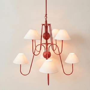 May include: A six-arm chandelier featuring a matte red metal frame with a central vertical rod and two spherical accents. The fixture holds six white conical fabric lampshades, each positioned at the end of a curved arm, suspended from a single rod.