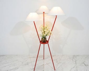 May include: A red metal floor lamp with three white lampshades. The lamp has a tripod base and a small round tray in the center holding a small potted plant with white flowers. The lamp is a decorative home decor item.