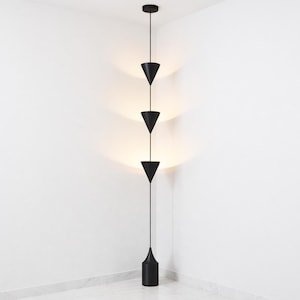 May include: A modern black pendant light fixture with three inverted cone-shaped shades suspended from a ceiling mount. The fixture extends downward to a cylindrical base. The light casts a warm glow against the white wall.