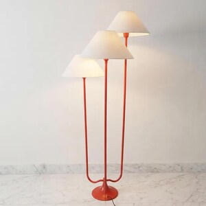 May include: A three-light floor lamp with a vibrant orange metal frame and off-white lampshades. The lamp features a round base and a unique, curved design. The lampshades are conical, and the lamp is positioned against a white wall.