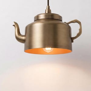 May include: A pendant light fixture shaped like a teapot with a brushed brass finish. The metal body features a curved spout and a handle on the side. A single light bulb is visible inside the open bottom, casting a warm glow downward.