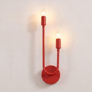 Pode incluir: Um candeeiro de parede vermelho brilhante com uma estrutura metálica em forma de U que suporta dois casquilhos verticais tipo vela. Cada casquilho contém uma lâmpada clara e brilhante, montada numa placa de base circular vermelha.
