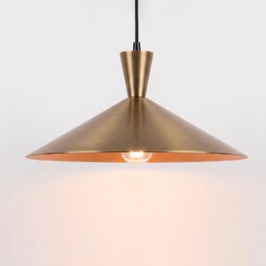 May include: A pendant light fixture featuring a wide, conical shade with a brushed brass finish. The shade is attached to a slender, matching brass neck and a black cord. A single light bulb is visible, emitting a warm glow from the center.