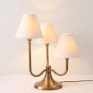 May include: A table lamp with a brass-toned metal base and three curved arms, each supporting a white conical fabric shade. The lamp features a central stem and two side stems of varying heights, all emitting a soft, warm glow from within.
