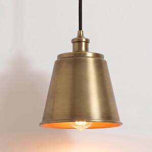 May include: A pendant light fixture featuring a bell-shaped shade with a brushed brass finish. The fixture is suspended by a thin black cord and includes a single exposed light bulb emitting a warm glow from within the metallic housing.