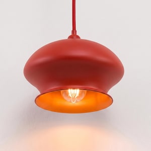 May include: A mushroom-shaped pendant light fixture with a matte red exterior finish and a warm, glowing orange interior. The lamp is suspended by a matching red cord and features a single exposed filament light bulb centered inside the shade.